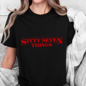 May include: Black t-shirt featuring the red text "SIXTY SEVEN THINGS". The text is in a bold, stylized font. The short-sleeved shirt is made of a soft material. The text is centered on the front of the shirt.