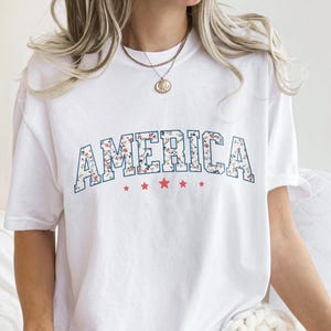 May include: White t-shirt featuring the word "AMERICA" in a floral pattern, outlined in blue, across the chest. Red stars are below the word. A casual, patriotic tee.