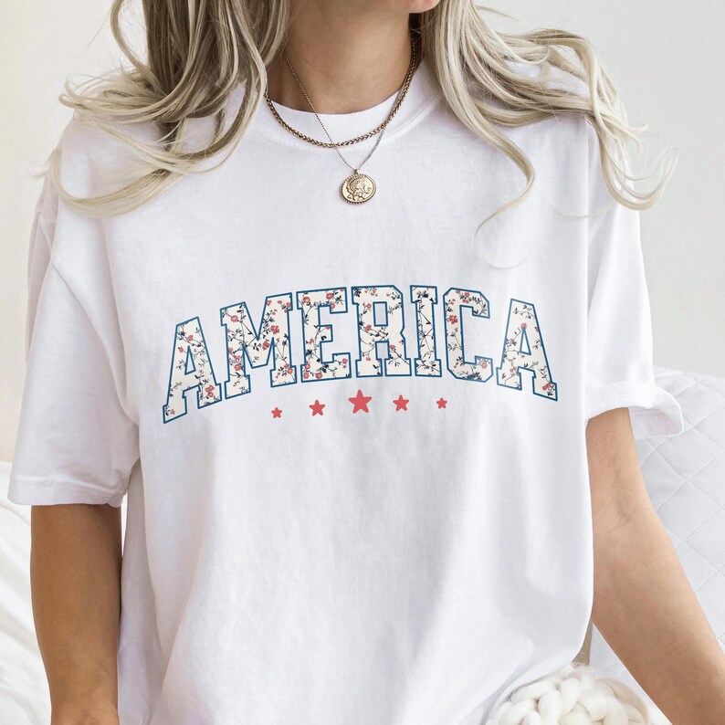 May include: White t-shirt featuring the word "AMERICA" in a floral pattern, outlined in blue, across the chest. Red stars are below the word. A casual, patriotic tee.