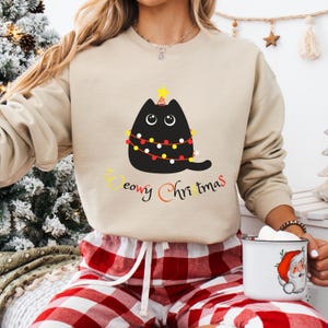 May include: A beige sweatshirt with a black cat wearing a Christmas lights garland and a star on its head. The text "Meowry Christmas" is printed below the cat.