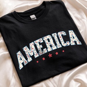 May include: Black t-shirt featuring the word "AMERICA" in a floral design with a light blue outline. Five red stars are below the text. The shirt is made of a soft material.