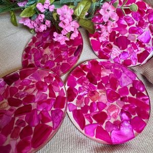 Puede incluir: Cuatro posavasos redondos con un diseño de mosaico de piedras rosas y magenta. Los posavasos tienen un borde dorado y están dispuestos sobre una superficie de color neutro. Flores rosas y hojas verdes en el fondo.