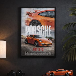 May include: Orange Porsche 911 GT3 poster in a black frame, displayed on a dark wood surface. A model car and a book with the word "PORSCHE" are also on the surface. The poster is 12"x18".