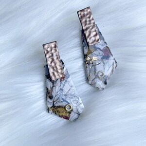 May include: Abstract earrings featuring a marbled design in white, grey, and burgundy, with gold leaf accents. Each earring has a hammered copper-coloured bar at the top and a small gold-coloured circle near the bottom.