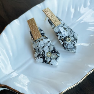 May include: Geometric earrings with a marbled pattern in black, white, and gold. Each earring has a gold-coloured rectangular accent at the top and a small, round, gold-coloured embellishment. The earrings are displayed on a white, leaf-shaped dish.