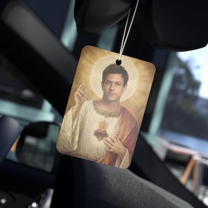 May include: Rectangular car air freshener featuring a portrait of a man in religious attire with a halo. The image has a vintage style, with the man's portrait in the center, surrounded by a golden glow. The air freshener hangs from a string.