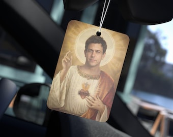 May include: Rectangular car air freshener featuring a portrait of a man in religious attire with a halo. The image has a vintage style, with the man's portrait in the center, surrounded by a golden glow. The air freshener hangs from a string.