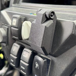 May include: A close-up of a black dashboard with various switches and controls. A black accessory is mounted on the dashboard. The dashboard has a textured surface and includes a radio and other electronic components. The "ROOF LIGHTS" switch is visible.