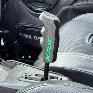 May include: A custom gray and black shift knob with the text "KRX" in green. The shift knob is installed in a vehicle with black seats and a Kawasaki speaker visible in the background. The text "T-REX OFF" is also visible.