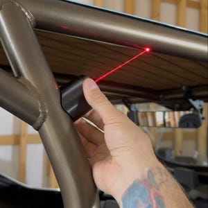 May include: A black cylindrical laser alignment tool mounted on a metallic bronze roll cage frame. The device emits a thin, straight red laser beam that projects onto the upper horizontal bar of the structure to assist with precise positioning.