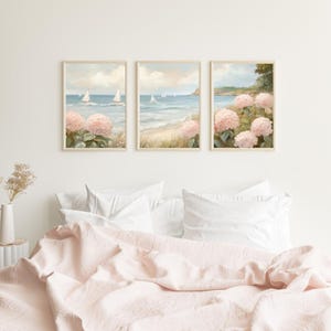 May include: Three framed wall art prints depicting a coastal scene with sailboats on the water, a sandy beach, and pink hydrangea flowers. The artwork has a soft, pastel color palette and is displayed above a bed with a pink comforter.