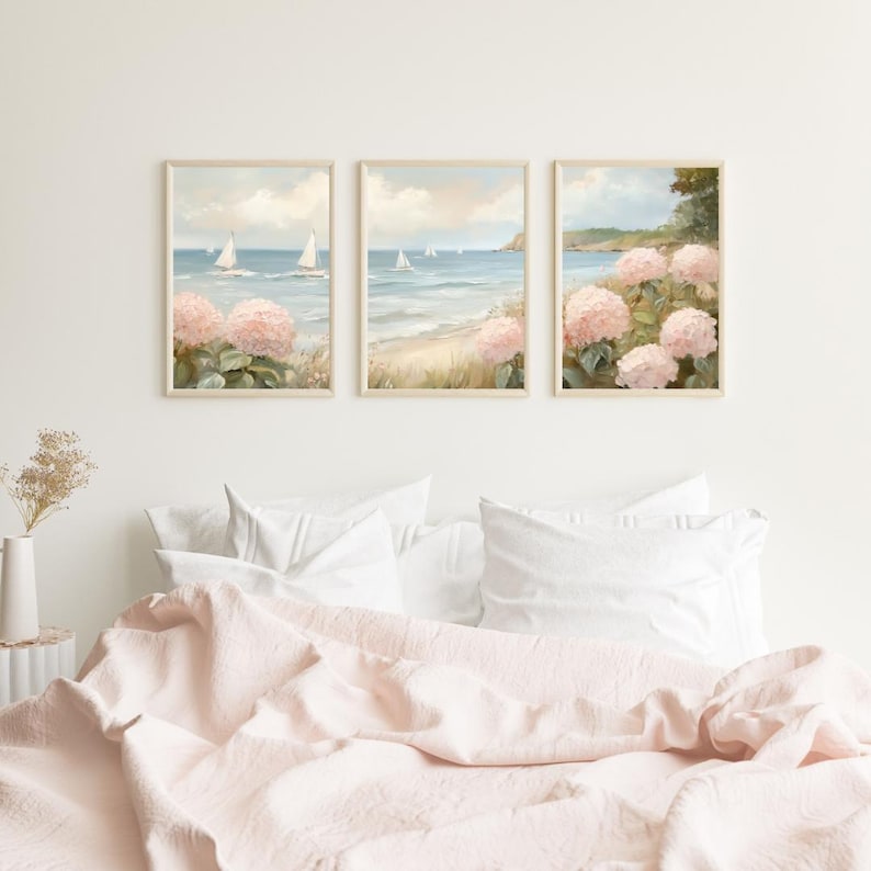 May include: Three framed wall art prints depicting a coastal scene with sailboats on the water, a sandy beach, and pink hydrangea flowers. The artwork has a soft, pastel color palette and is displayed above a bed with a pink comforter.
