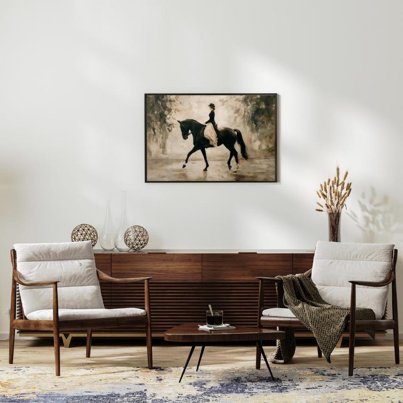 May include: A framed black and white equestrian art print depicting a rider on a black horse. The print hangs above a wooden cabinet, flanked by two armchairs with cream-colored cushions. A small, dark wood coffee table sits in front of the chairs.