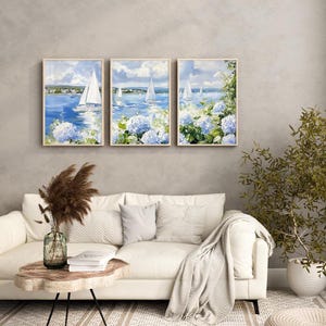 May include: Three framed wall art pieces depict sailboats on a blue sea under a cloudy sky. The paintings feature white sailboats, blue hydrangeas, and a coastal landscape. The artwork is displayed above a white sofa with decorative pillows.