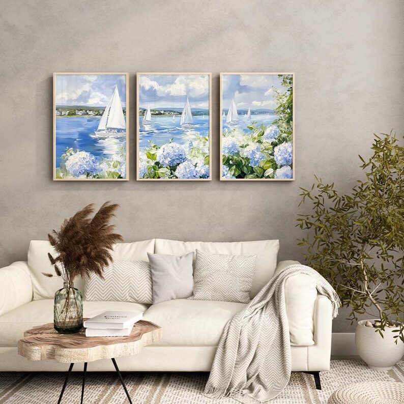 May include: Three framed wall art pieces depict sailboats on a blue sea under a cloudy sky. The paintings feature white sailboats, blue hydrangeas, and a coastal landscape. The artwork is displayed above a white sofa with decorative pillows.