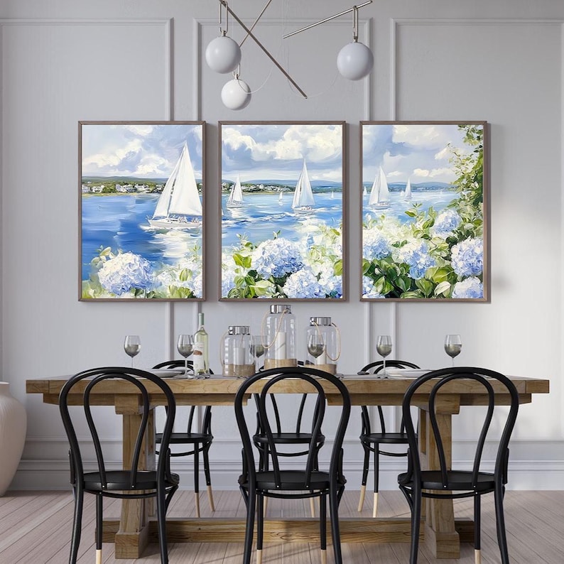 May include: A triptych of framed paintings depicting sailboats on a blue sea under a cloudy sky. The paintings are surrounded by blue hydrangeas. The paintings are displayed above a wooden dining table with black chairs.