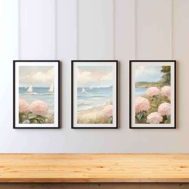 May include: Set of three framed art prints. Each print depicts a coastal scene with sailboats on the water, pink hydrangeas, and a sandy beach. The artwork is framed in black and hangs on a white wall.