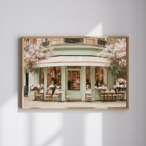 K&ouml;nnte beinhalten: Ein gerahmter Druck eines Pariser Caf&eacute;s mit einer mintgr&uuml;nen Fassade, einer rosa Markise und Sitzgelegenheiten im Freien. Das Caf&eacute; ist mit rosa Blumen geschm&uuml;ckt und verf&uuml;gt &uuml;ber mehrere kleine Tische und St&uuml;hle im Freien. Das Bild weckt ein Gef&uuml;hl von europ&auml;ischem Charme.