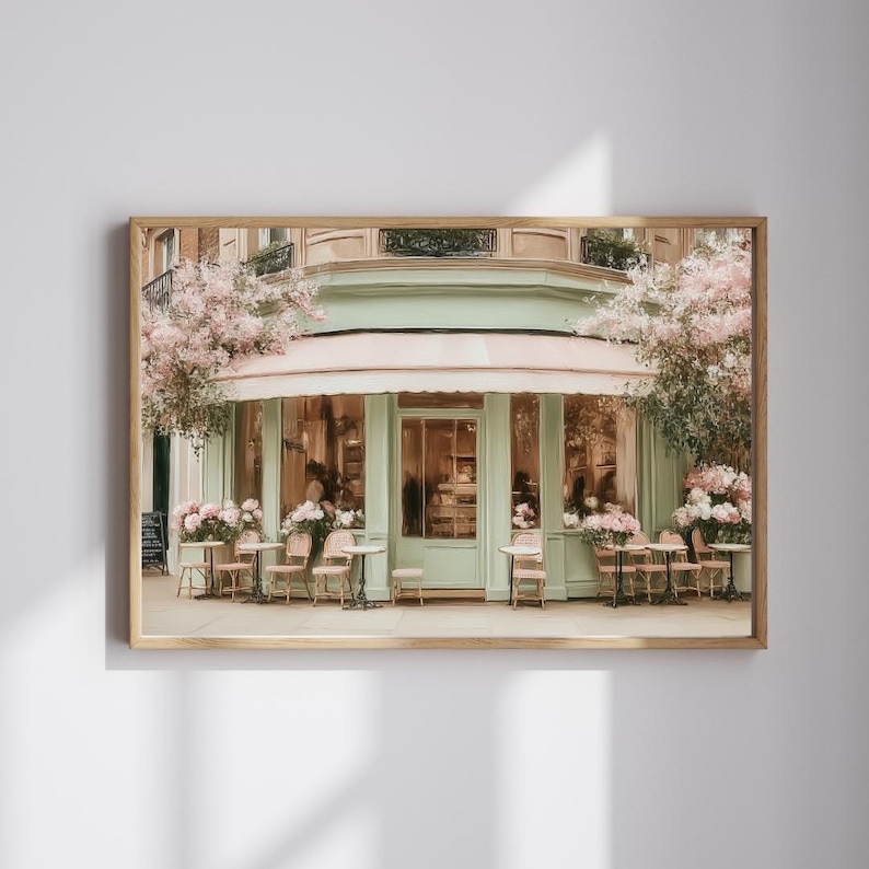 K&ouml;nnte beinhalten: Ein gerahmter Druck eines Pariser Caf&eacute;s mit einer mintgr&uuml;nen Fassade, einer rosa Markise und Sitzgelegenheiten im Freien. Das Caf&eacute; ist mit rosa Blumen geschm&uuml;ckt und verf&uuml;gt &uuml;ber mehrere kleine Tische und St&uuml;hle im Freien. Das Bild weckt ein Gef&uuml;hl von europ&auml;ischem Charme.