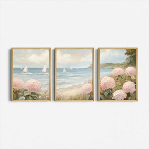 May include: Set of three framed art prints. Each print features a coastal scene with sailboats on the water, a sandy beach, and pink hydrangea flowers. The frames are a light wood color, and the overall style is soft and inviting.