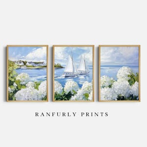 Puede incluir: Conjunto de tres láminas artísticas enmarcadas con escenas costeras. La obra representa veleros en el agua, flores blancas y casas junto a la orilla. Las láminas están enmarcadas en madera. El texto "RANFURLY PRINTS" está en la parte inferior.