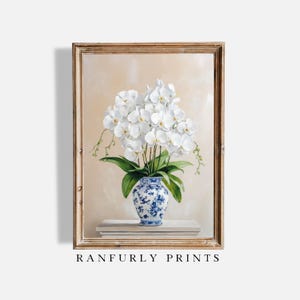 Op de afbeelding: Een ingelijst schilderij van witte orchideeën in een blauw-witte vaas. De vaas staat op een stapel boeken. De schildering heeft een lichtbeige achtergrond. De lijst is lichtbruin van kleur. De tekst "RANFURLY PRINTS" staat onderaan de afbeelding.