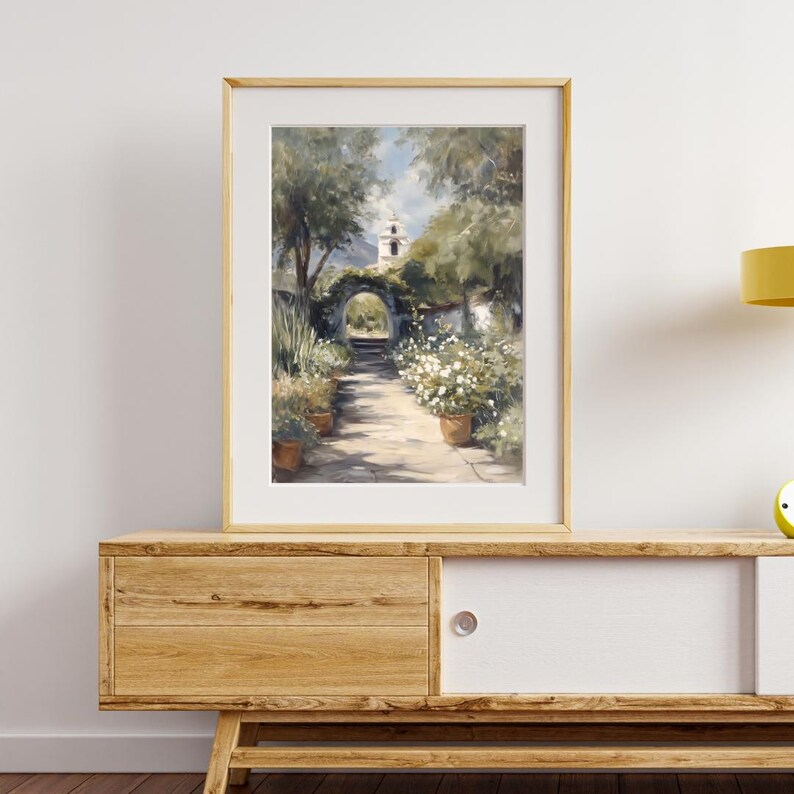 以下が含まれることがあります： 日当たりの良い庭の小道がアーチ型の入り口と遠くの白い建物へと続く、額装されたアートプリント。アートワークは、緑豊かな緑、テラコッタの鉢植えの白い花、明るい色のフレームが特徴です。