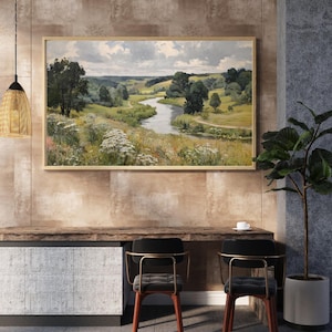 May include: A framed landscape painting depicts a winding river through a green valley under a cloudy sky. The artwork is displayed above a wooden desk with two chairs. A decorative light fixture hangs from the ceiling, and a potted plant is on the right.