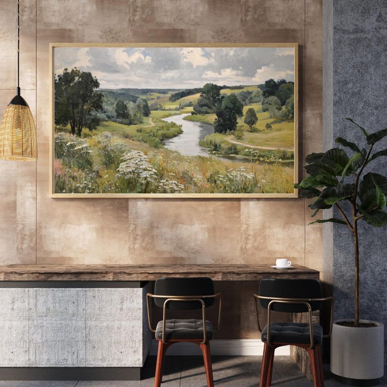 May include: A framed landscape painting depicts a winding river through a green valley under a cloudy sky. The artwork is displayed above a wooden desk with two chairs. A decorative light fixture hangs from the ceiling, and a potted plant is on the right.