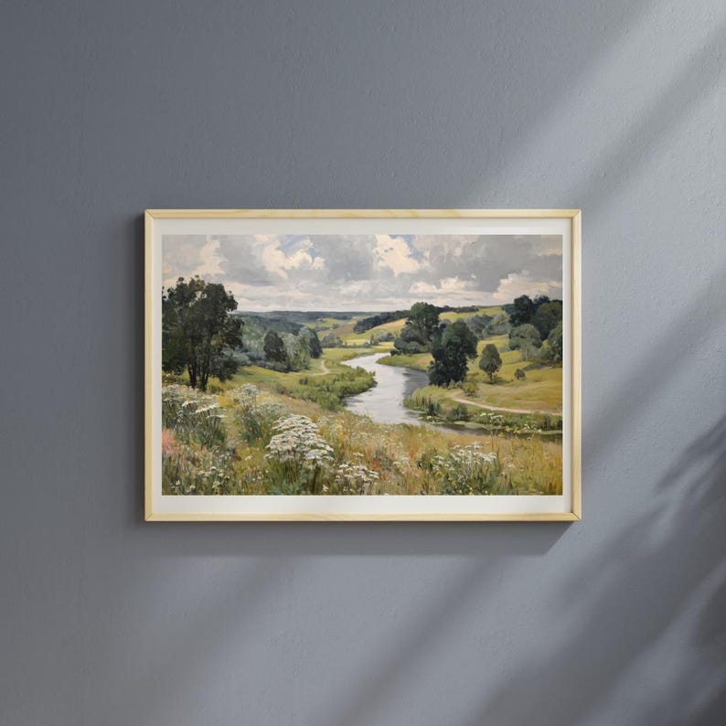 May include: A framed landscape painting depicts a serene river winding through a valley. The artwork features lush green fields, trees, and a cloudy sky. The painting is encased in a light-colored wooden frame, creating a classic aesthetic.
