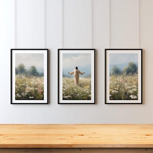 May include: Three framed art prints depicting a figure in a field of wildflowers with arms outstretched. The prints have black frames and are hung on a white wall above a wooden table. The artwork features a serene landscape with a soft, natural color palette.