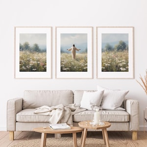 May include: Three framed art prints depicting a figure in a field of wildflowers with arms outstretched. The prints have light wood frames and are displayed above a beige sofa with wooden side tables. The artwork has a soft, pastel color palette.