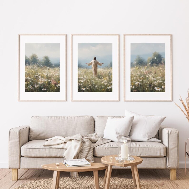 May include: Three framed art prints depicting a figure in a field of wildflowers with arms outstretched. The prints have light wood frames and are displayed above a beige sofa with wooden side tables. The artwork has a soft, pastel color palette.