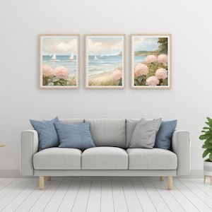 May include: Three framed landscape art prints featuring coastal scenes with sailboats, pink hydrangeas, and a beach. The prints are displayed above a light gray sofa with blue and gray throw pillows.