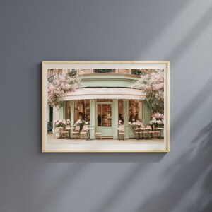 K&ouml;nnte beinhalten: Gerahmter Kunstdruck eines Pariser Caf&eacute;s mit mintgr&uuml;ner Fassade und rosa Markise. Tische und St&uuml;hle stehen drau&szlig;en, umgeben von rosa Blumen. Das Bild vermittelt einen Hauch von europ&auml;ischem Charme und Eleganz.