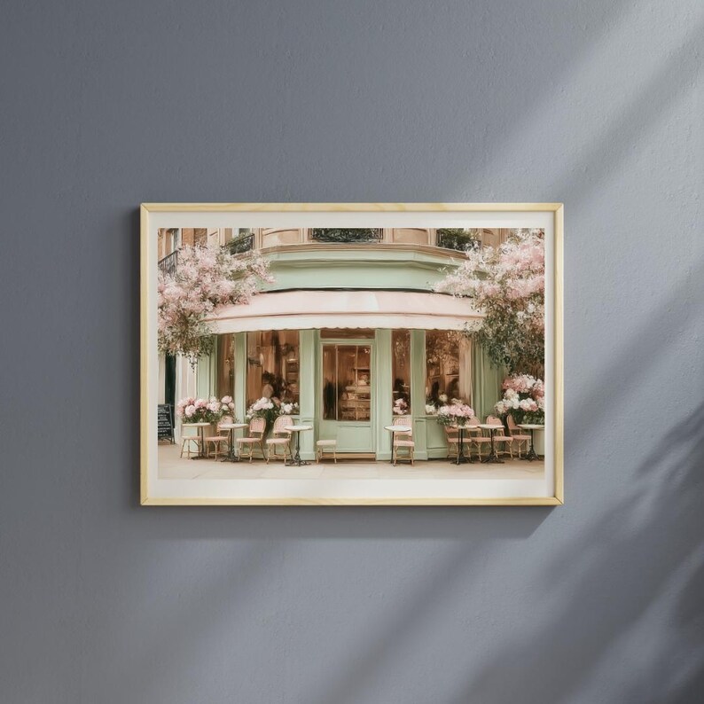 K&ouml;nnte beinhalten: Gerahmter Kunstdruck eines Pariser Caf&eacute;s mit mintgr&uuml;ner Fassade und rosa Markise. Tische und St&uuml;hle stehen drau&szlig;en, umgeben von rosa Blumen. Das Bild vermittelt einen Hauch von europ&auml;ischem Charme und Eleganz.