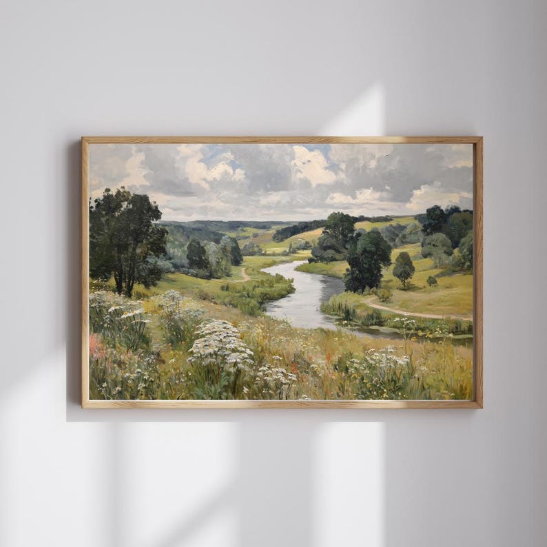 May include: A framed landscape painting depicts a serene river winding through a lush valley. The artwork features a palette of greens, yellows, and blues, with trees, fields, and a cloudy sky. The painting is in a light-colored wooden frame.