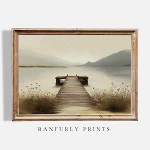 Op de afbeelding: Een ingelijste landschapsafdruk met een houten pier die zich uitstrekt in een kalm meer, met bergen op de achtergrond. Het kunstwerk heeft een gedempt kleurenpalet van beige, grijs en bruin. De tekst "RANFURLY PRINTS" staat onderaan.