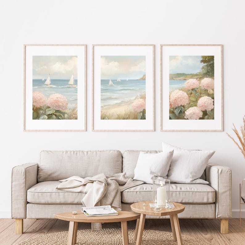 May include: Three framed landscape art prints. Each print depicts a coastal scene with sailboats on the water, a sandy beach, and pink hydrangea flowers. The artwork has a soft, pastel color palette with a light wooden frame.