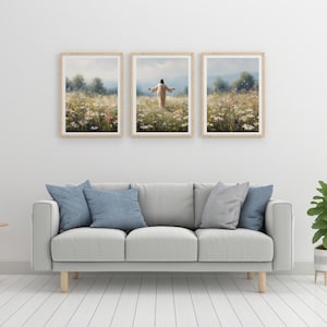 May include: Three framed landscape art prints depicting a person in a field of wildflowers. The prints have light wood frames and are hung above a gray sofa with blue pillows. The artwork features a person with arms outstretched.