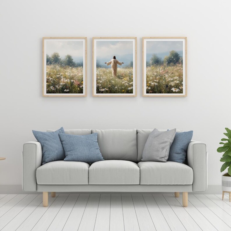 May include: Three framed landscape art prints depicting a person in a field of wildflowers. The prints have light wood frames and are hung above a gray sofa with blue pillows. The artwork features a person with arms outstretched.