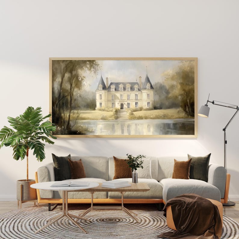 K&ouml;nnte beinhalten: Ein gerahmtes Aquarellbild eines wei&szlig;en Schlosses mit einem grauen Dach, umgeben von B&auml;umen und einem reflektierenden Teich. Das Bild h&auml;ngt an einer wei&szlig;en Wand &uuml;ber einem hellgrauen Ecksofa mit braunen Kissen.