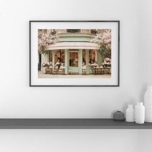 K&ouml;nnte beinhalten: Ein gerahmter Druck eines Pariser Caf&eacute;s mit einer mintgr&uuml;nen Markise und Fassade. Tische und St&uuml;hle sind drau&szlig;en aufgestellt, mit rosa Blumen und Gr&uuml;n. Das Bild vermittelt einen Hauch von europ&auml;ischem Charme und Eleganz.