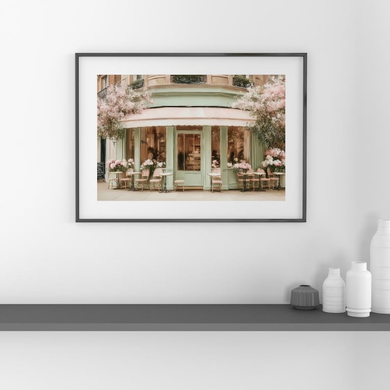 K&ouml;nnte beinhalten: Ein gerahmter Druck eines Pariser Caf&eacute;s mit einer mintgr&uuml;nen Markise und Fassade. Tische und St&uuml;hle sind drau&szlig;en aufgestellt, mit rosa Blumen und Gr&uuml;n. Das Bild vermittelt einen Hauch von europ&auml;ischem Charme und Eleganz.