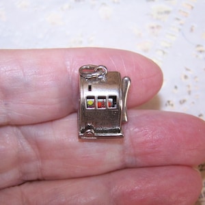 May include: A small silver-toned metal charm shaped like a vintage slot machine. The charm features a side lever, a top jump ring for attachment, and three small windows displaying colored symbols including a yellow circle and a red circle.