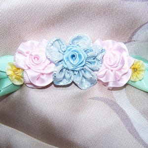 May include: A floral hair accessory featuring three fabric flowers in pink and blue, with green leaves and yellow accents. The flowers are arranged in a row, creating a decorative and delicate design.