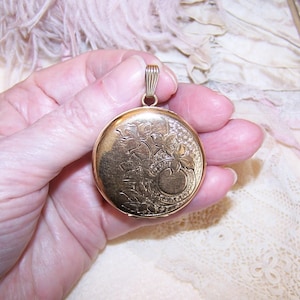 May include: A gold-colored round locket with a floral design. The locket has a detailed engraved pattern of flowers and leaves. It features a small loop at the top for a chain. The locket is held in a hand.