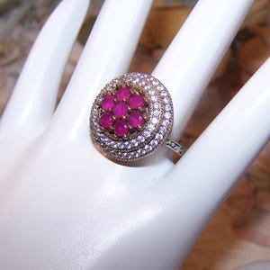 May include: A silver ring with a cluster of small, round, bright pink gemstones at the center, surrounded by a halo of smaller, clear stones. The ring is set on a white surface.