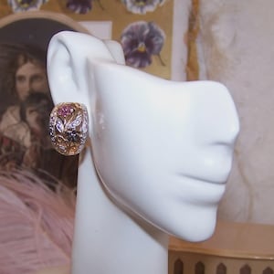 May include: A gold and silver-toned earring with a floral design, featuring pink, purple, and black gemstones. The earring is displayed on a white mannequin head, showcasing its intricate details and craftsmanship. The jewelry piece is a statement accessory.