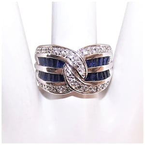 May include: A silver ring featuring a woven design. The ring is adorned with rows of dark blue gemstones and small, clear stones. The gemstones are set in a band around the ring's exterior. The ring is displayed on a white surface.